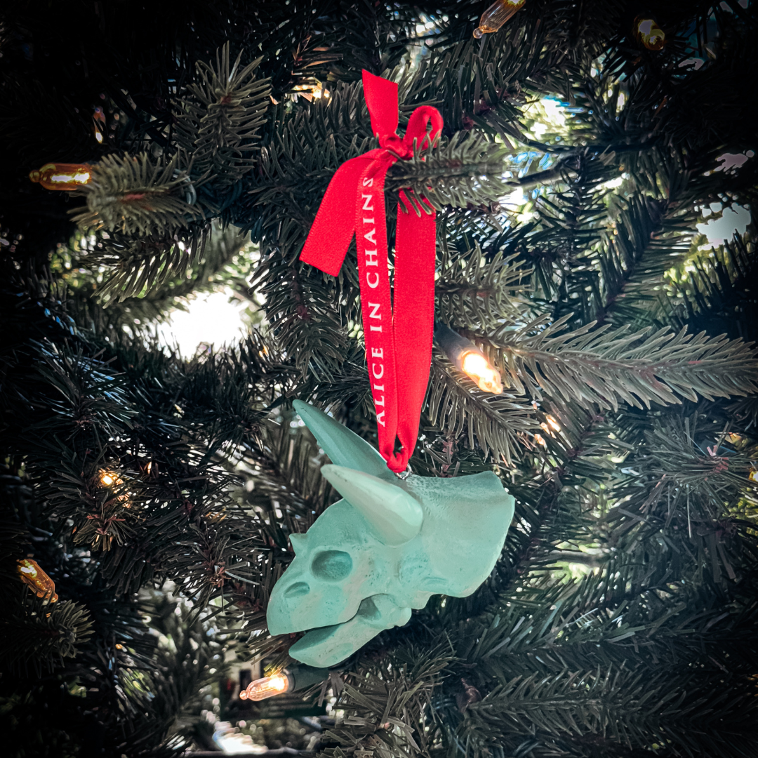 A mint-green triceratops skull holiday ornament with a red alice in chains ribbon in a holiday ornament box set